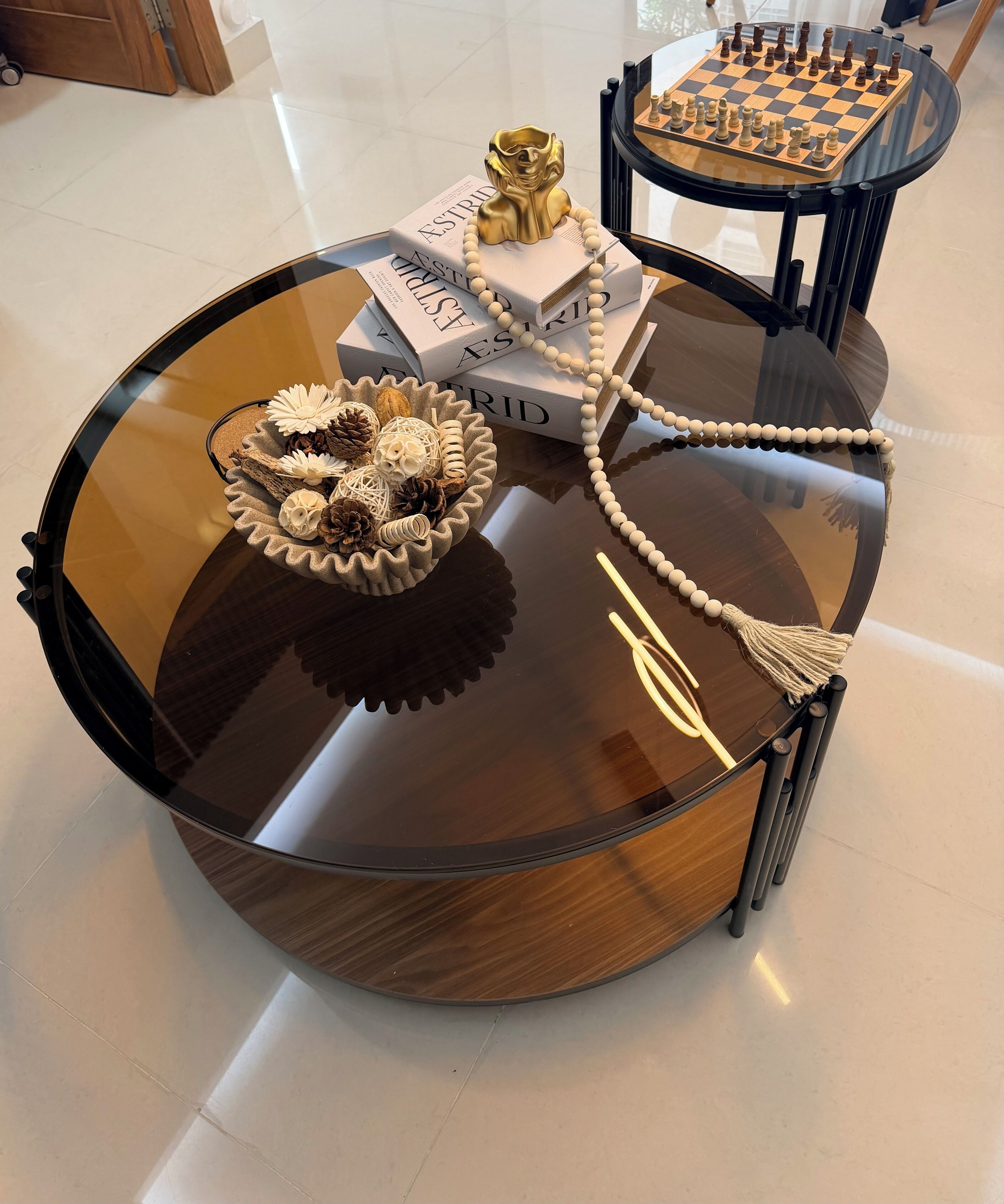 Une table ronde en verre, sur laquelle on trouve un jeu d’échecs, des livres, un objet doré et un collier de perles.