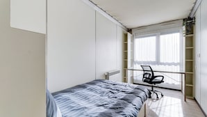 Ein Schlafzimmer mit Bett, Schreibtisch und Stuhl. Ein Fenster mit Jalousien und eine Heizung sind ebenfalls vorhanden.