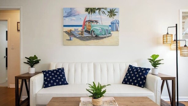 A living room with a white sofa, two side tables with plants, a coffee table with a tray and plant, and a beach-themed painting on the wall.