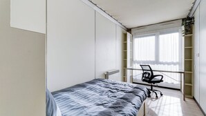 Un dormitorio con cama, escritorio y silla. Hay una ventana con persianas y un radiador.