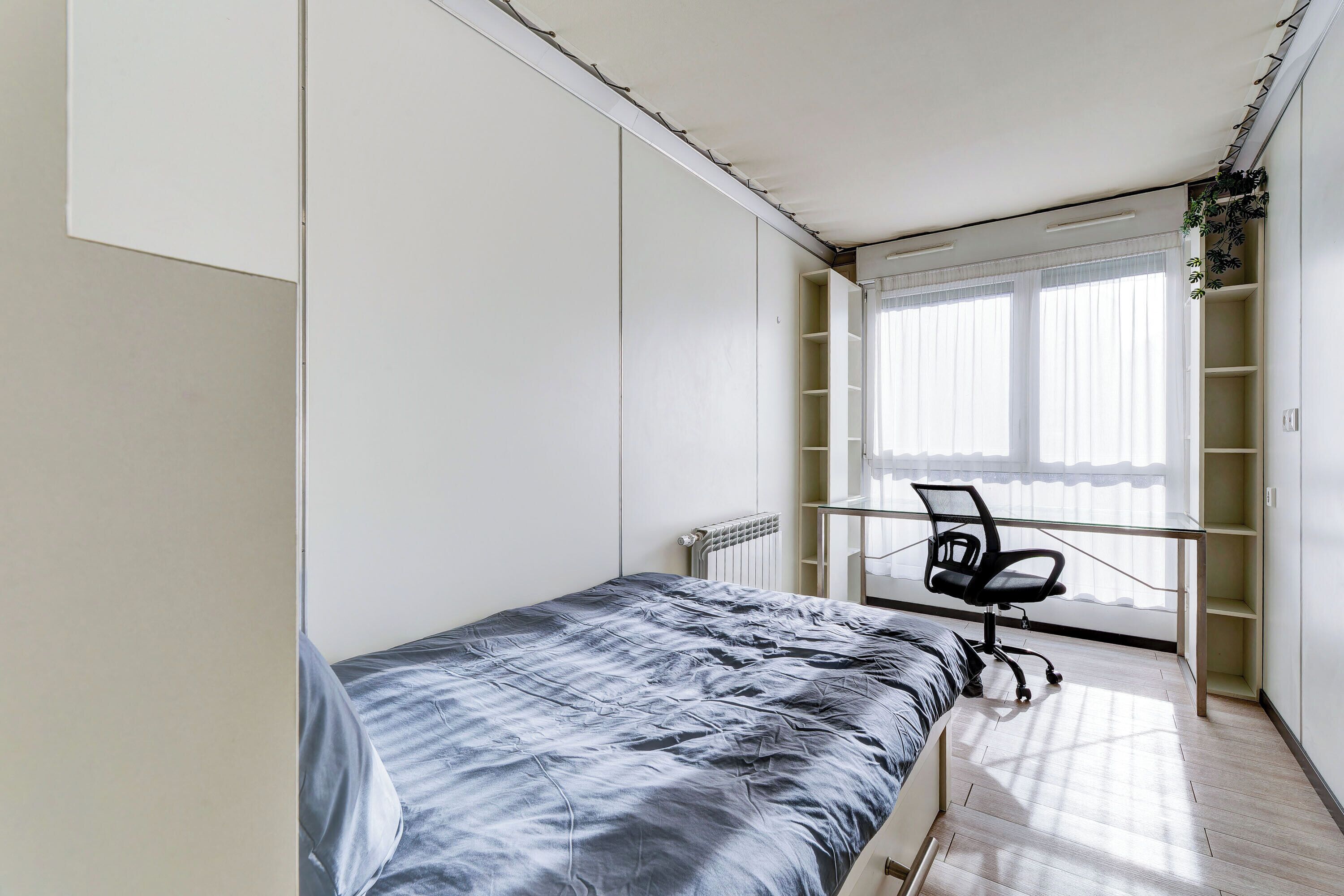 Ein Schlafzimmer mit Bett, Schreibtisch und Stuhl. Ein Fenster mit Jalousien und eine Heizung sind ebenfalls vorhanden.