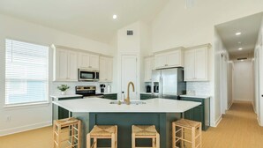 Una cocina moderna con armarios blancos, una isla central con taburetes, electrodomésticos de acero inoxidable y un ventanal con persianas.
