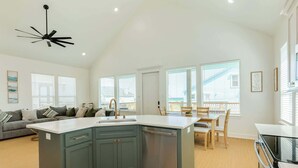 Una cocina moderna con una isla central, un comedor con mesa y sillas de madera, y una sala de estar con sofá y ventilador de techo.