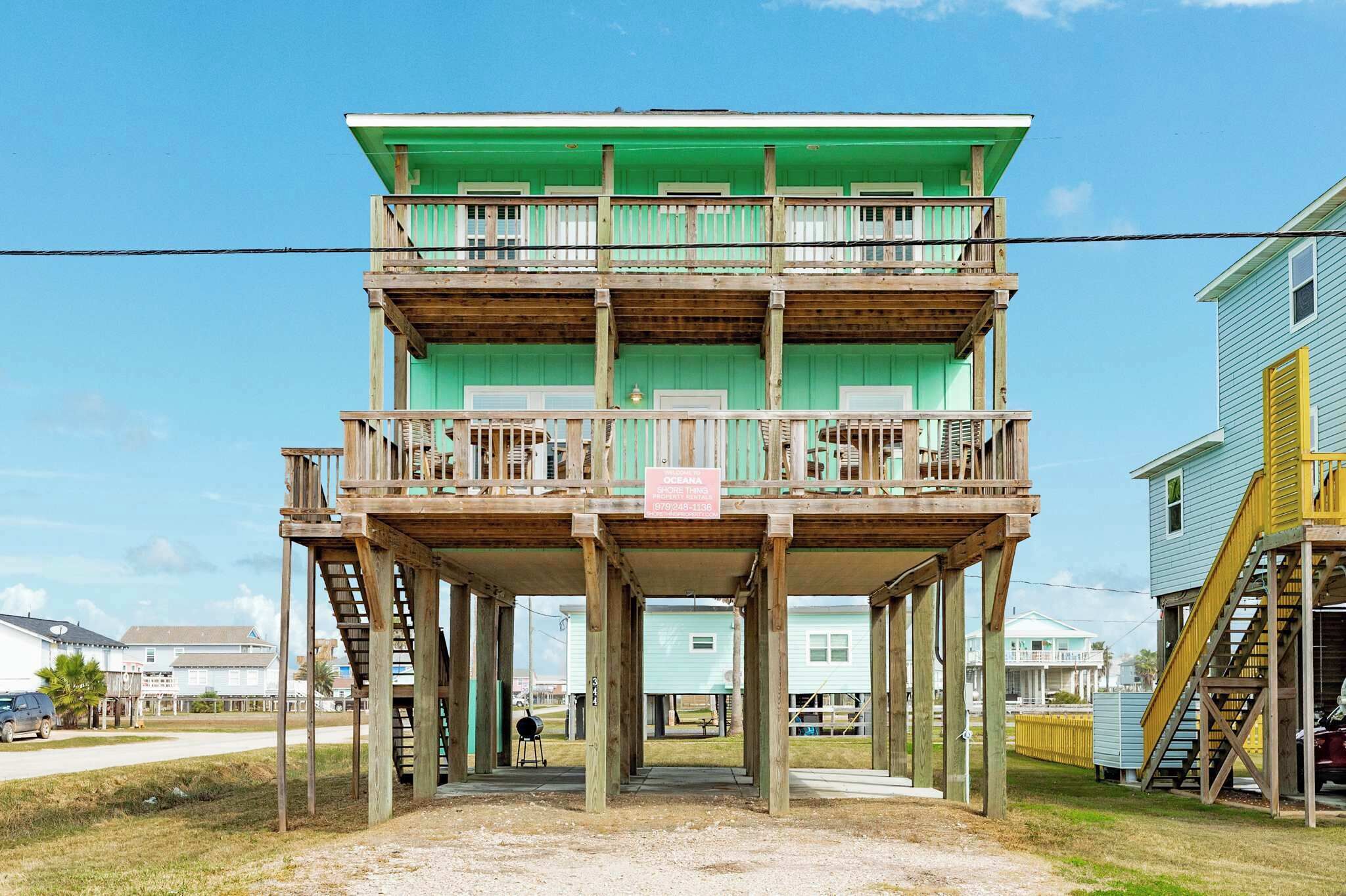 A multi-story wooden structure with a balcony, located on stilts over a sandy area.