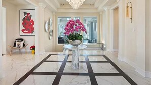 A spacious hallway with a marble floor, a central table with a vase of pink flowers, and a large chandelier. - Luxury Mansion available with Pool, Gym and More - Perfect for entertaining (Atlanta)
