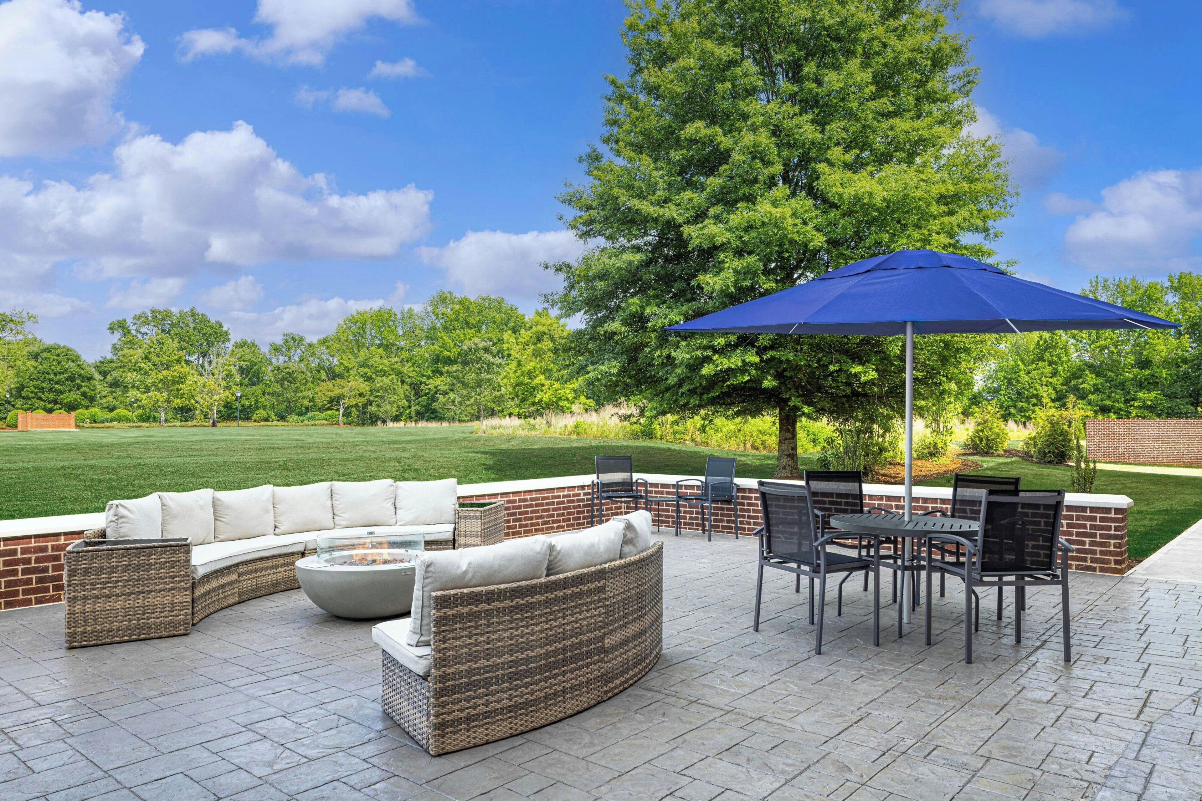 Een terras buiten met een gebogen bank, een eettafel met stoelen en een blauwe parasol.