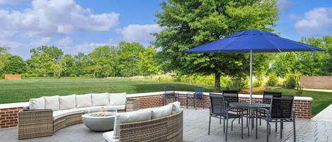 Een terras buiten met een gebogen bank, een eettafel met stoelen en een blauwe parasol.