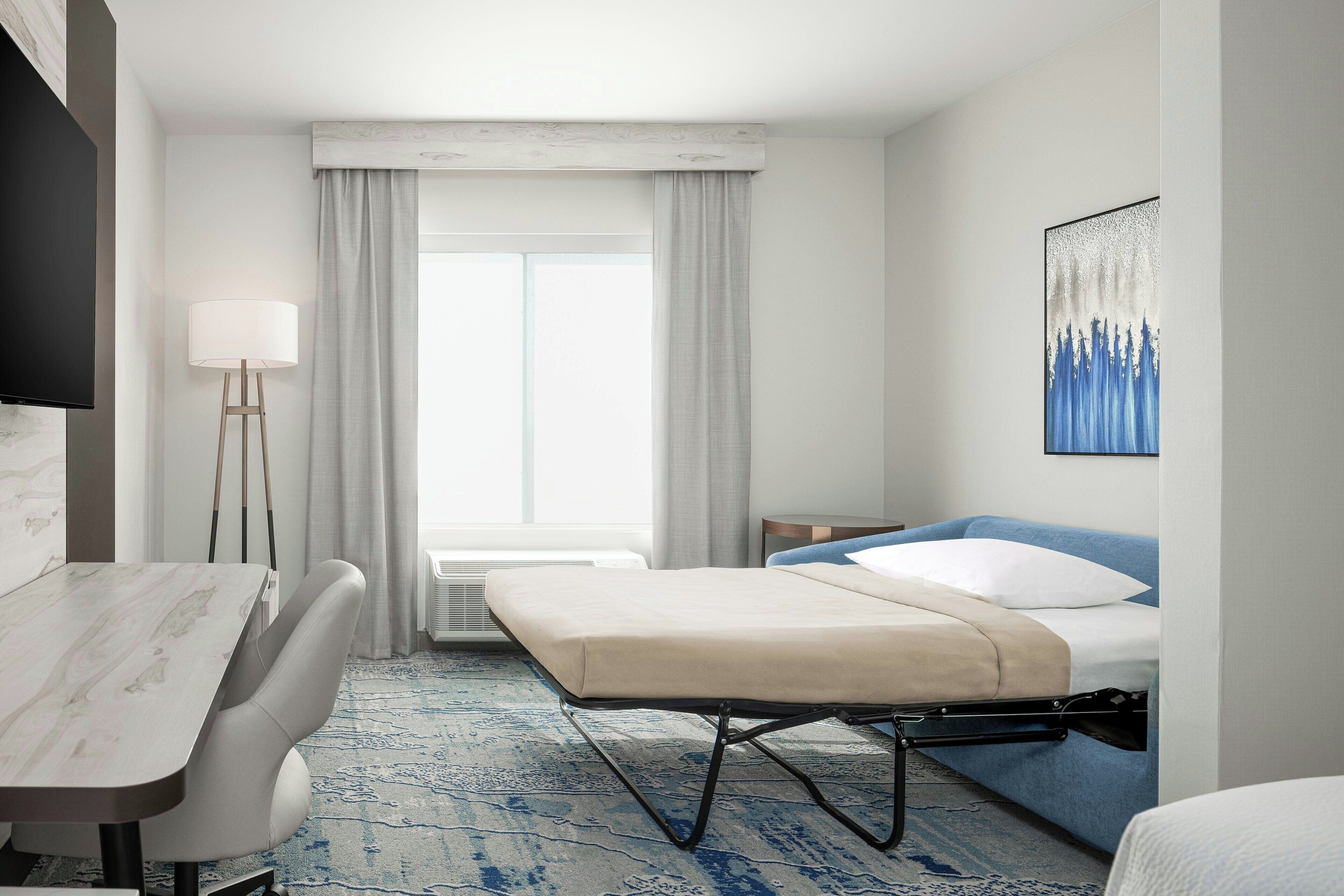 Habitación de hotel con cama, escritorio, silla, lámpara y un cuadro en la pared.