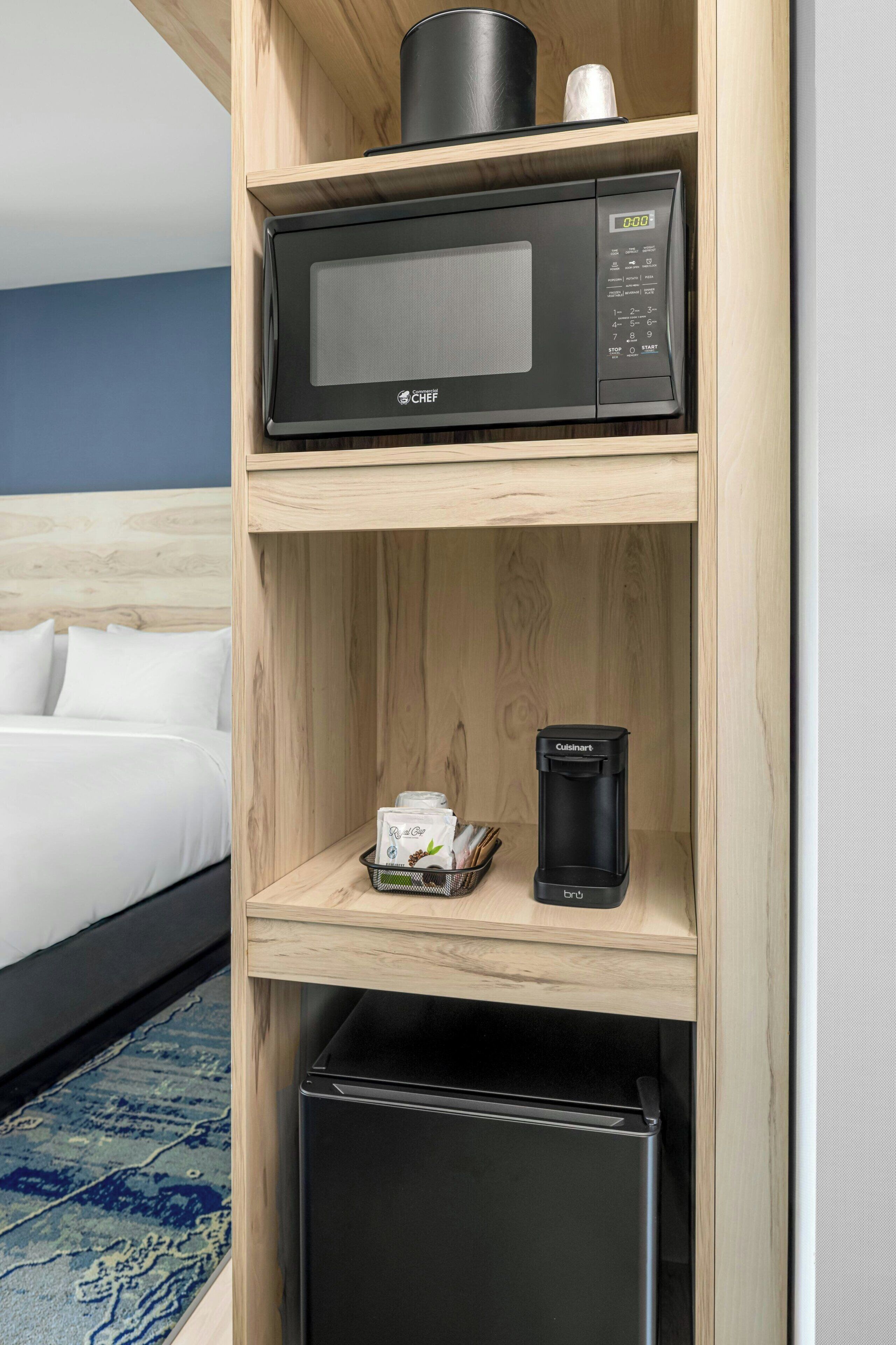 A hotel room with a microwave on the top shelf, a coffee maker on the middle shelf, and a refrigerator on the bottom shelf.