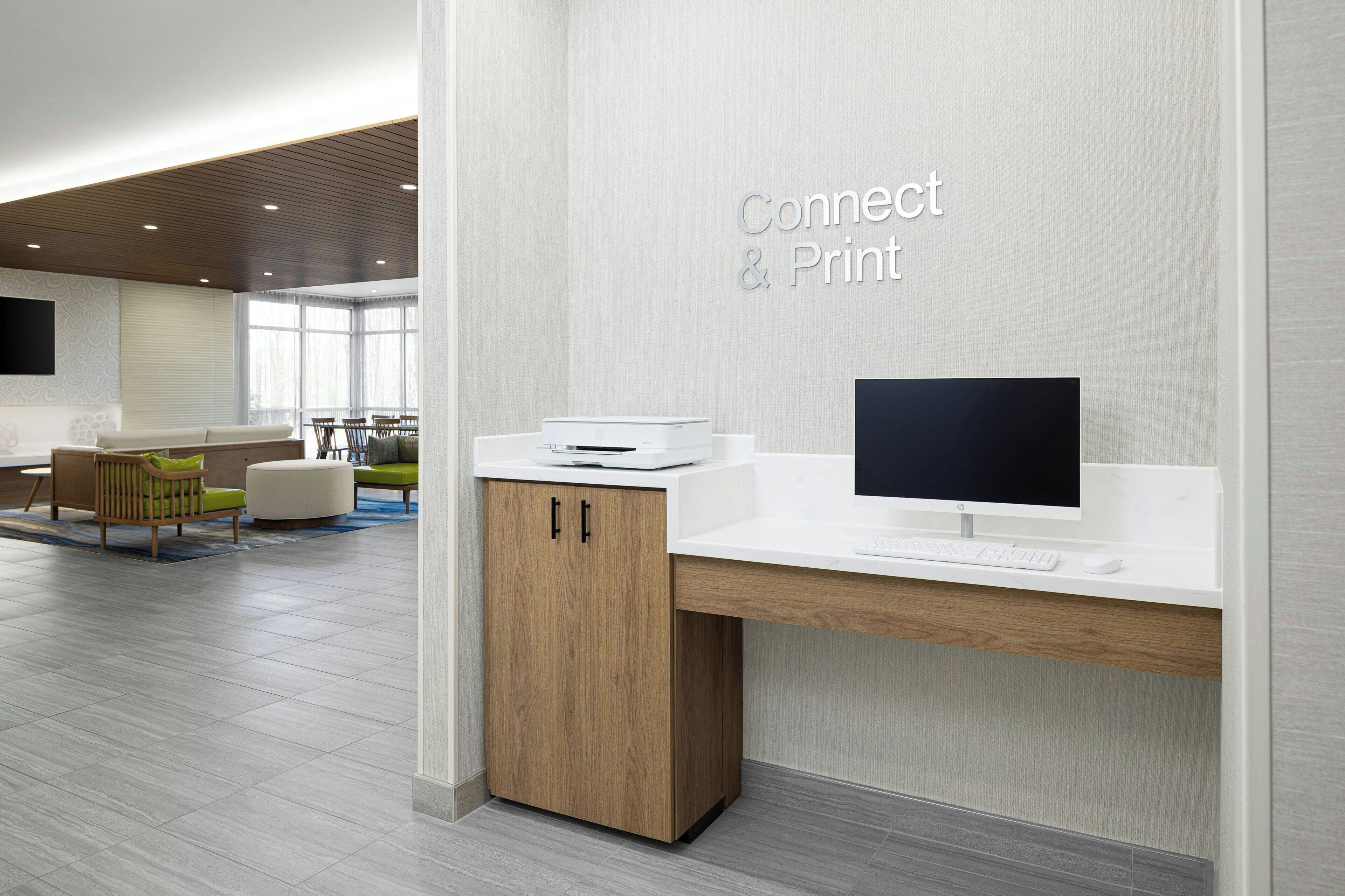 Un espace de bureau moderne avec un bureau blanc, un ordinateur et une imprimante. On aperçoit à l’arrière-plan un coin salon avec des chaises et une table.
