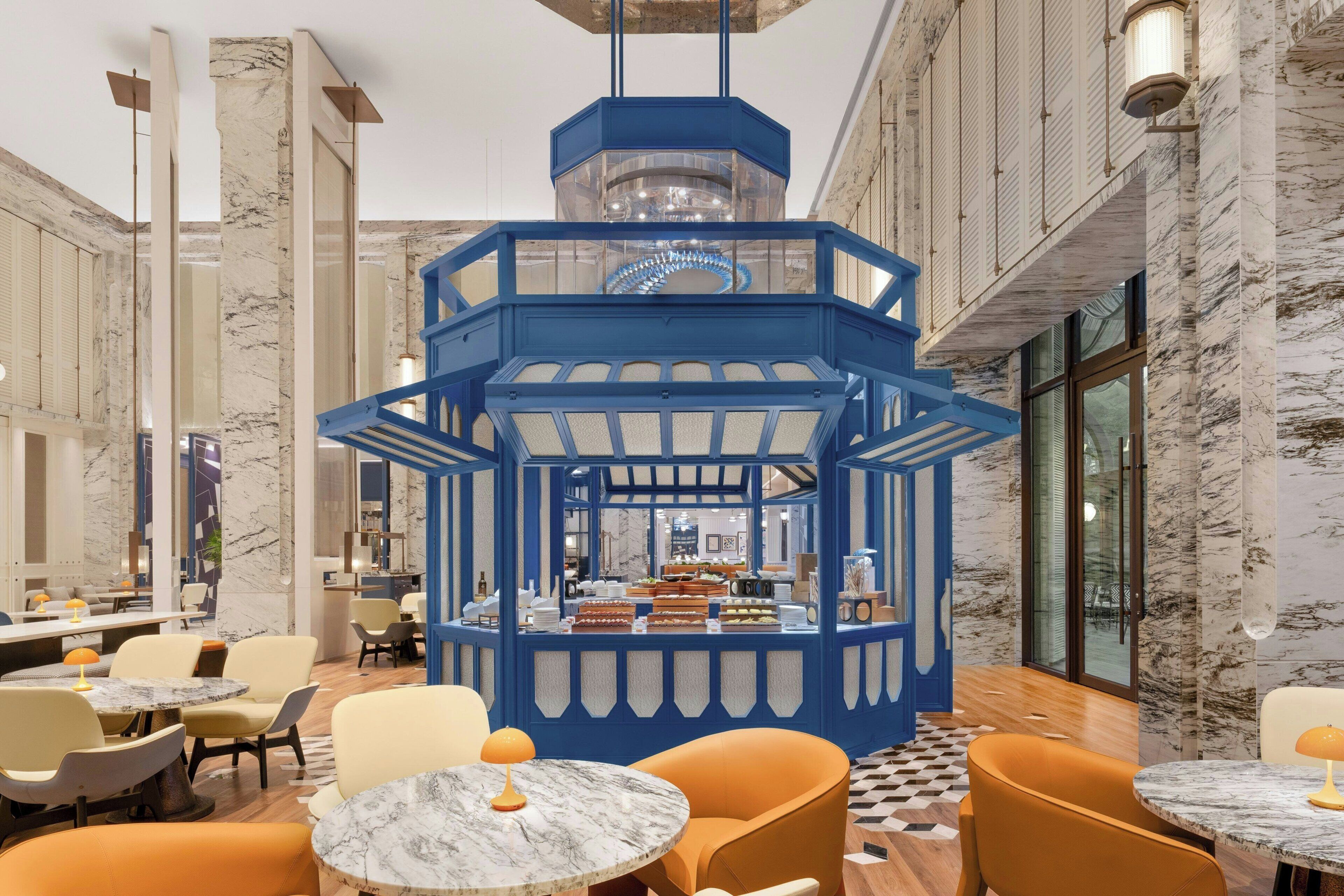 A hotel lobby with marble columns, a central blue kiosk, and seating areas with round tables.