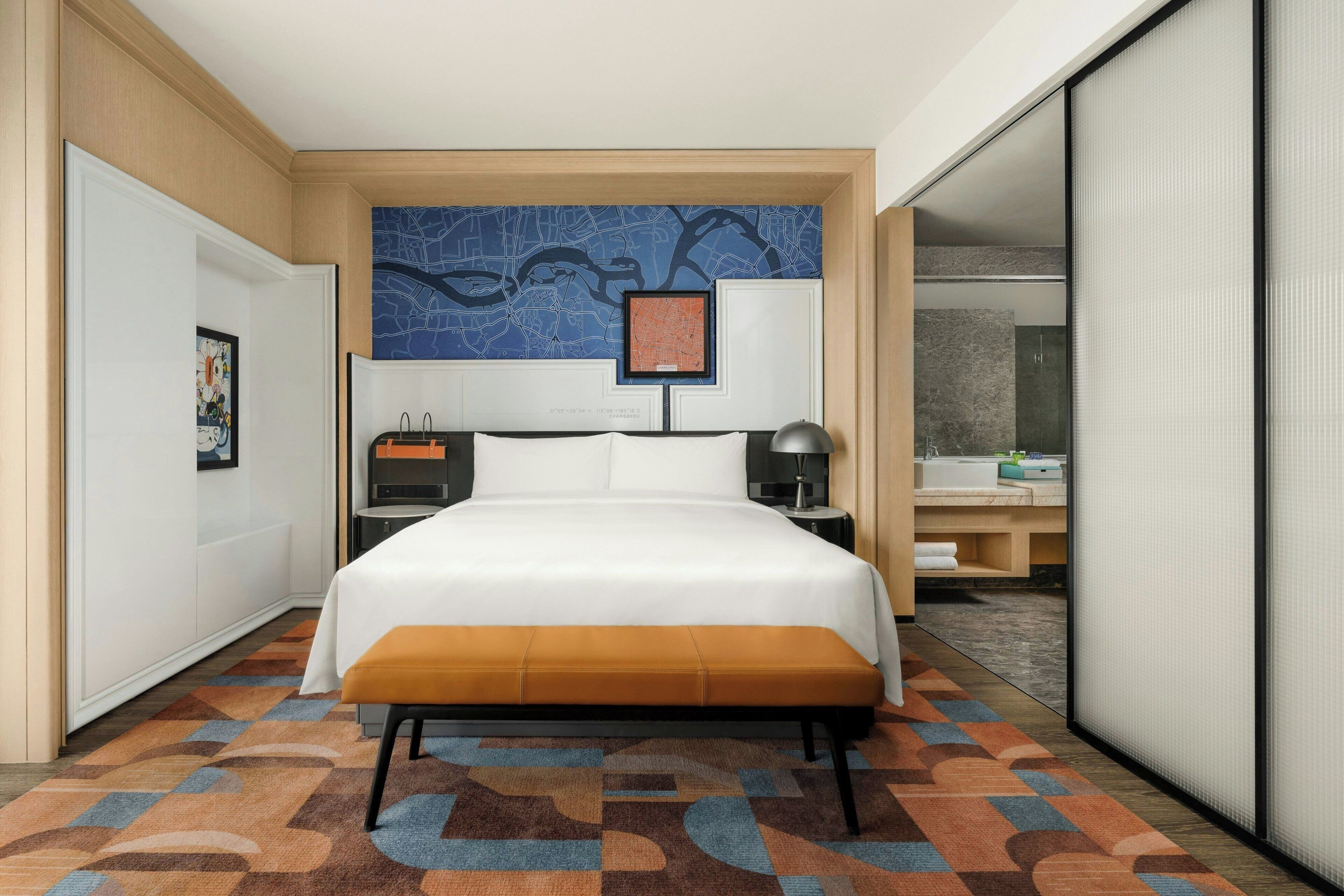 Habitación de hotel con una cama grande, un escritorio con lámpara, un mapa en la pared y un baño visible a través de una puerta abierta.