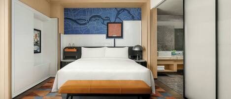 Habitación de hotel con una cama grande, un escritorio con lámpara, un mapa en la pared y un baño visible a través de una puerta abierta.