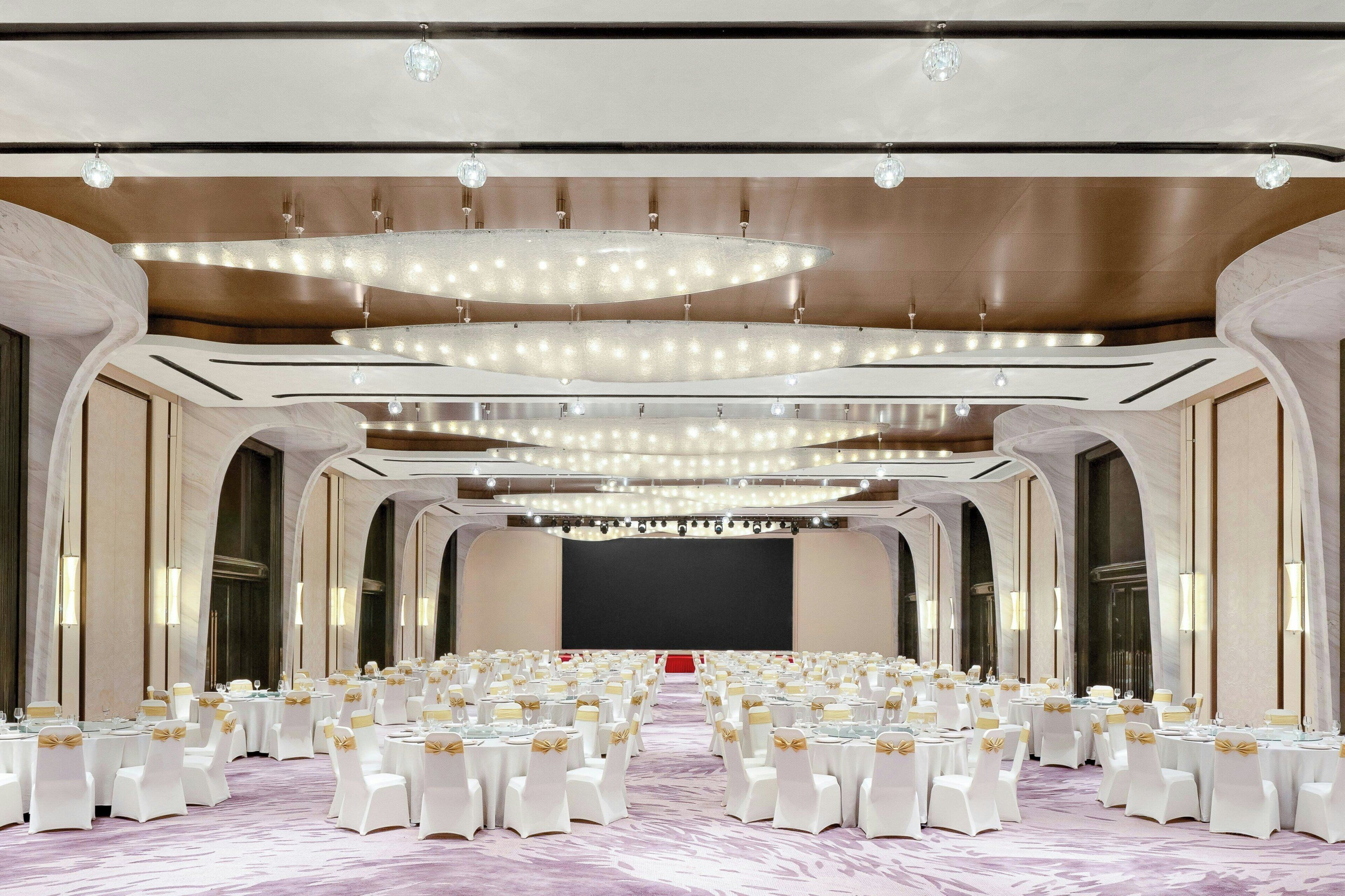 A banquet hall with round tables, white chairs, and chandeliers.
