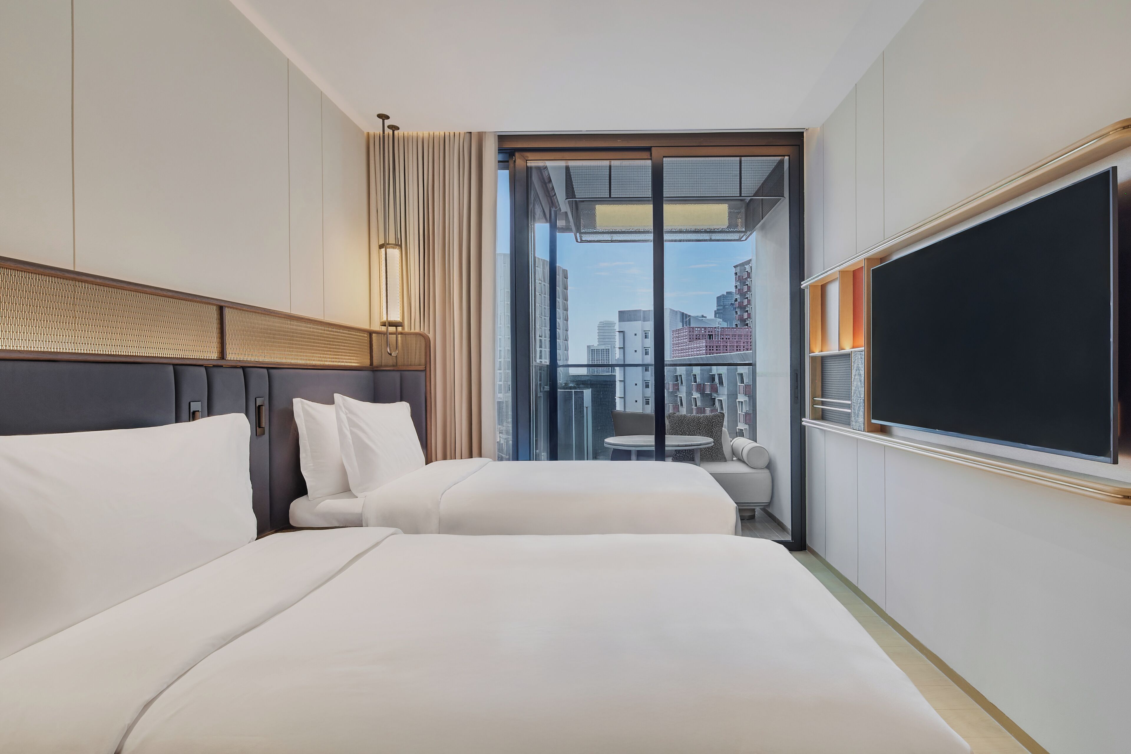 A hotel room with two beds, a large window with a city view, a flat-screen TV, and a small table.