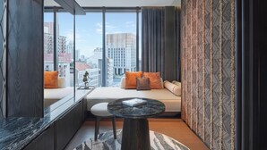 Een moderne hotelkamer met een groot raam, een bank met kussens, een klein rond tafeltje en een kamer met een patroon op de muur.