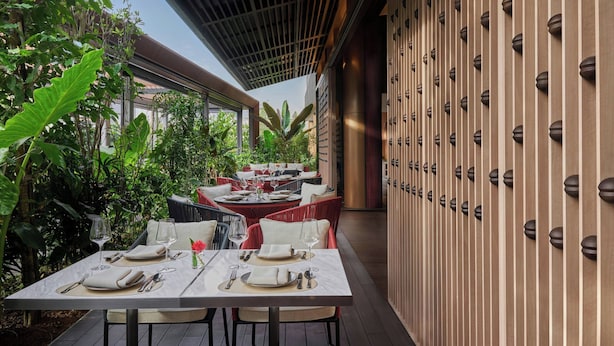 An outdoor dining area with tables set for two, surrounded by lush greenery and a wooden wall with decorative elements.