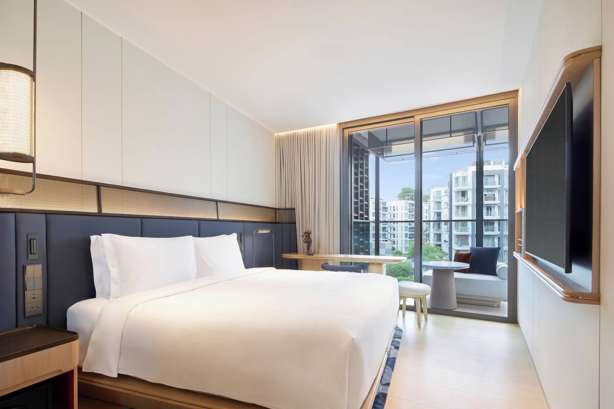 Habitación de hotel con cama, mesita de noche, lámpara y un ventanal con vistas a edificios.