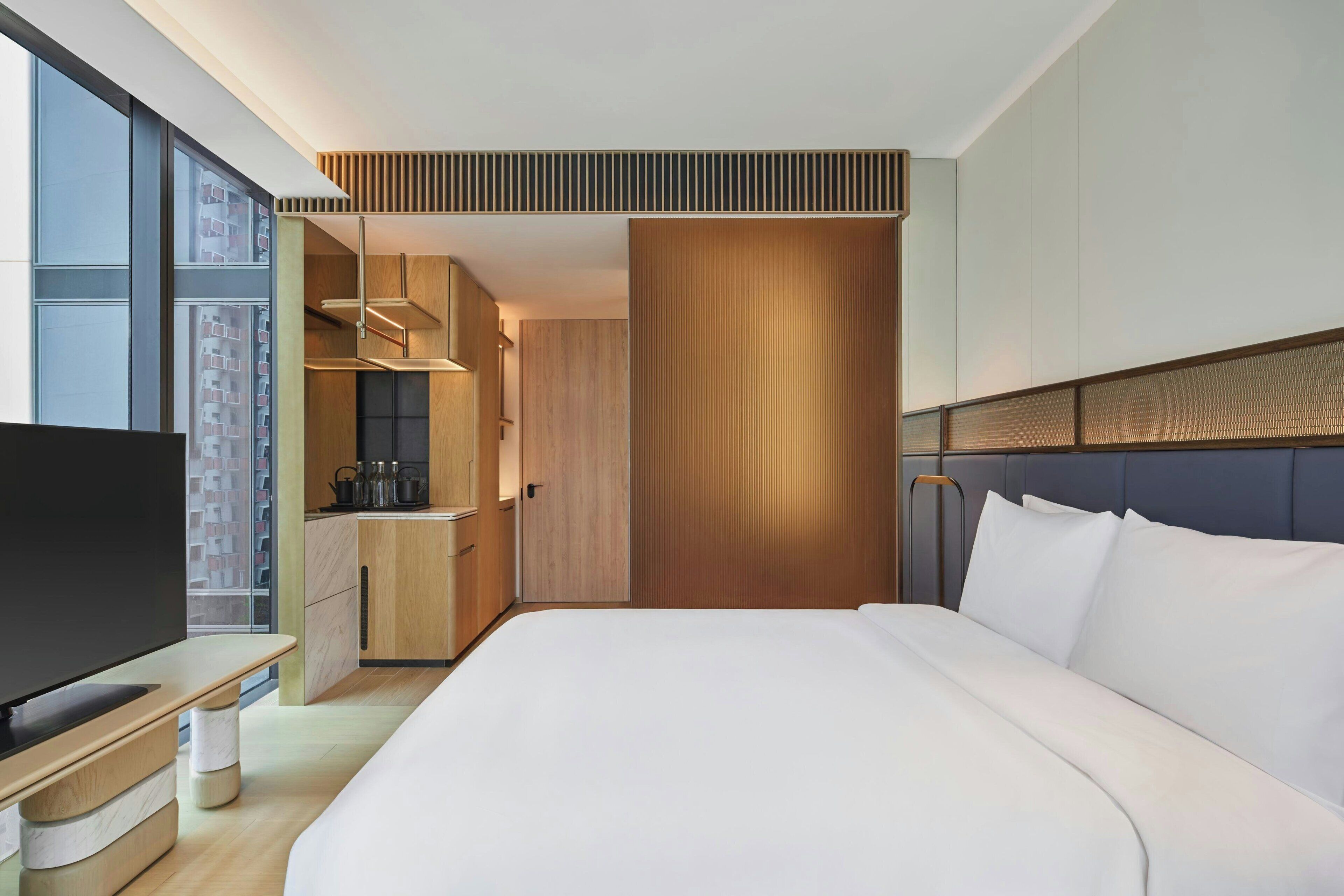 A modern hotel room with a large bed, a TV, a kitchenette, and a view of the city.