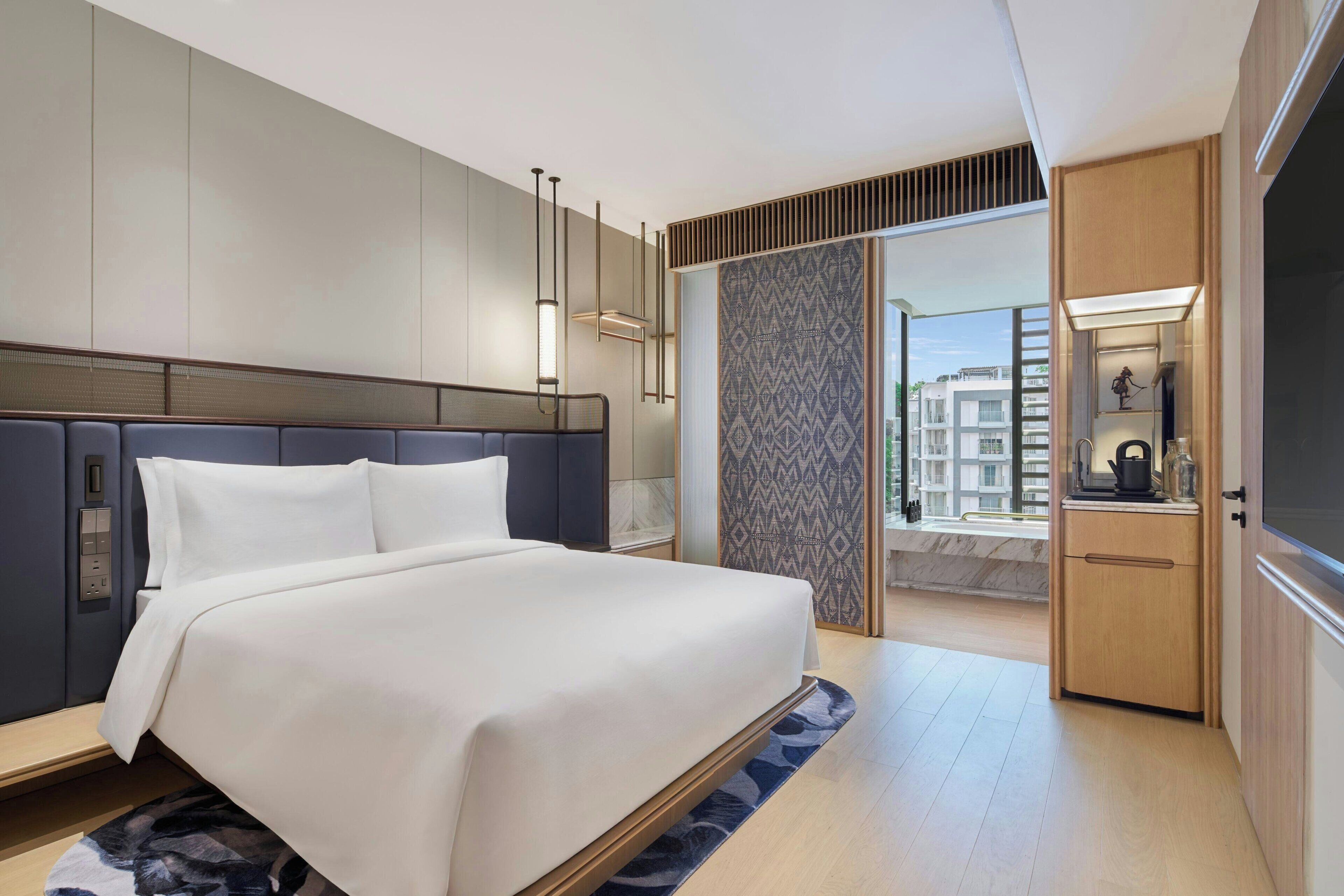 Ein modernes Hotelzimmer mit einem großen Bett, einer integrierten Garderobe, einer kleinen Küchenzeile und Blick auf die Stadtlandschaft.