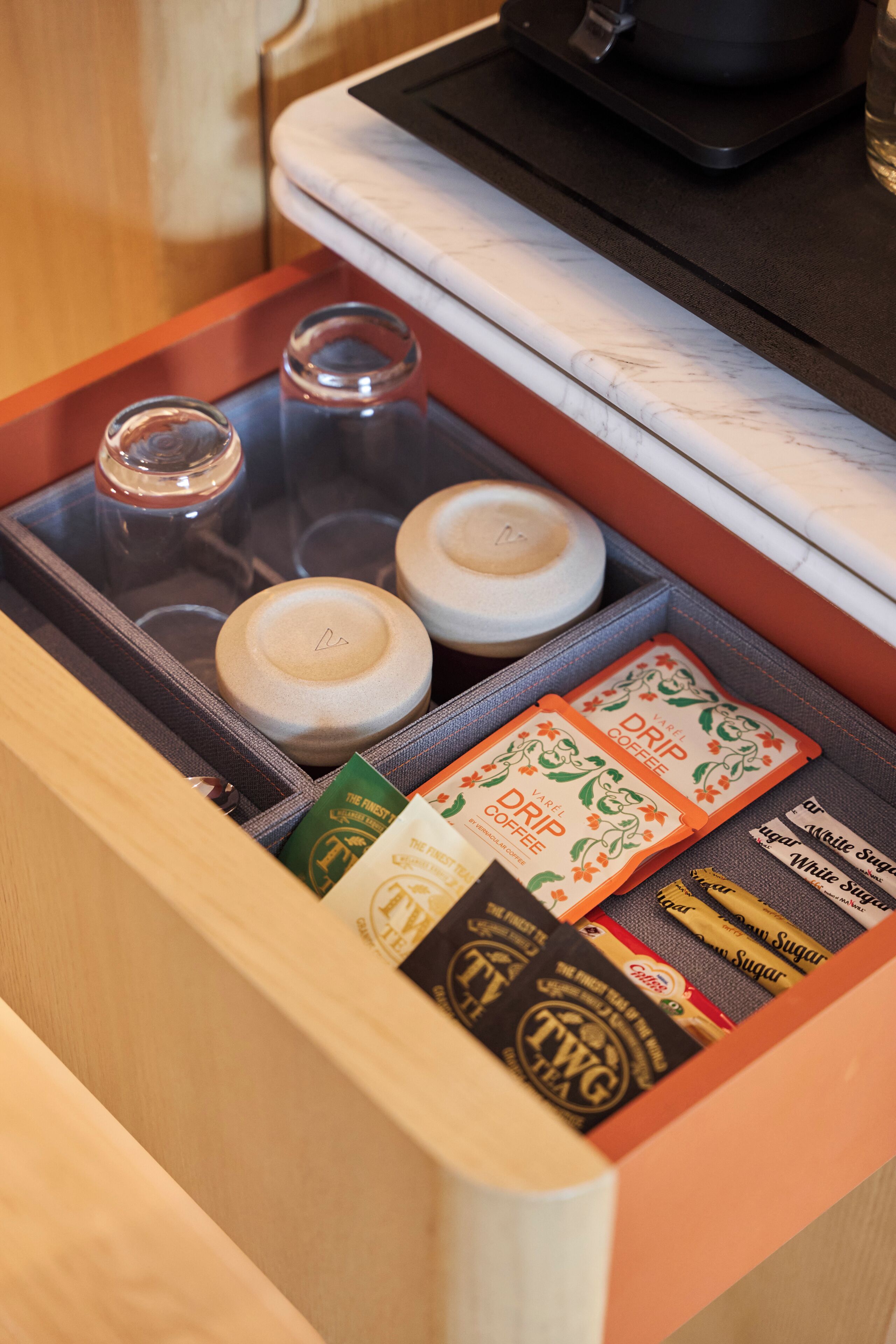 Un cajón con diversos objetos, incluyendo frascos, una cafetera y sobres de café.