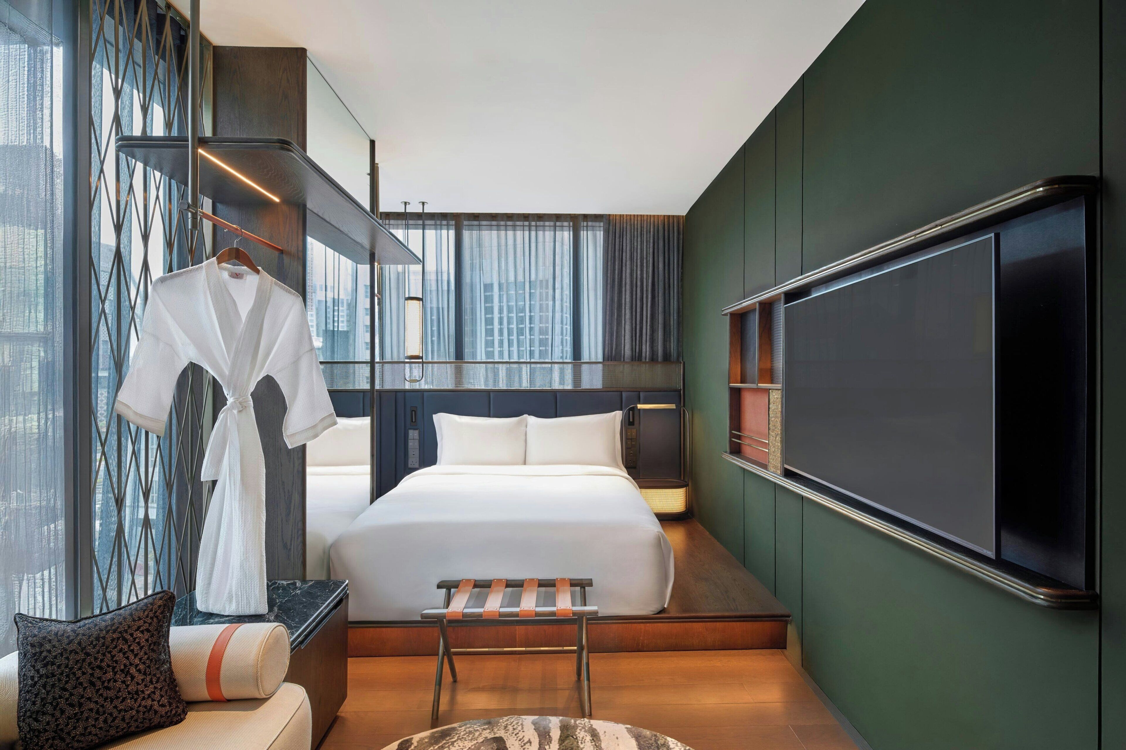 A modern hotel room with a large bed, a TV, a seating area, and a view of the city.