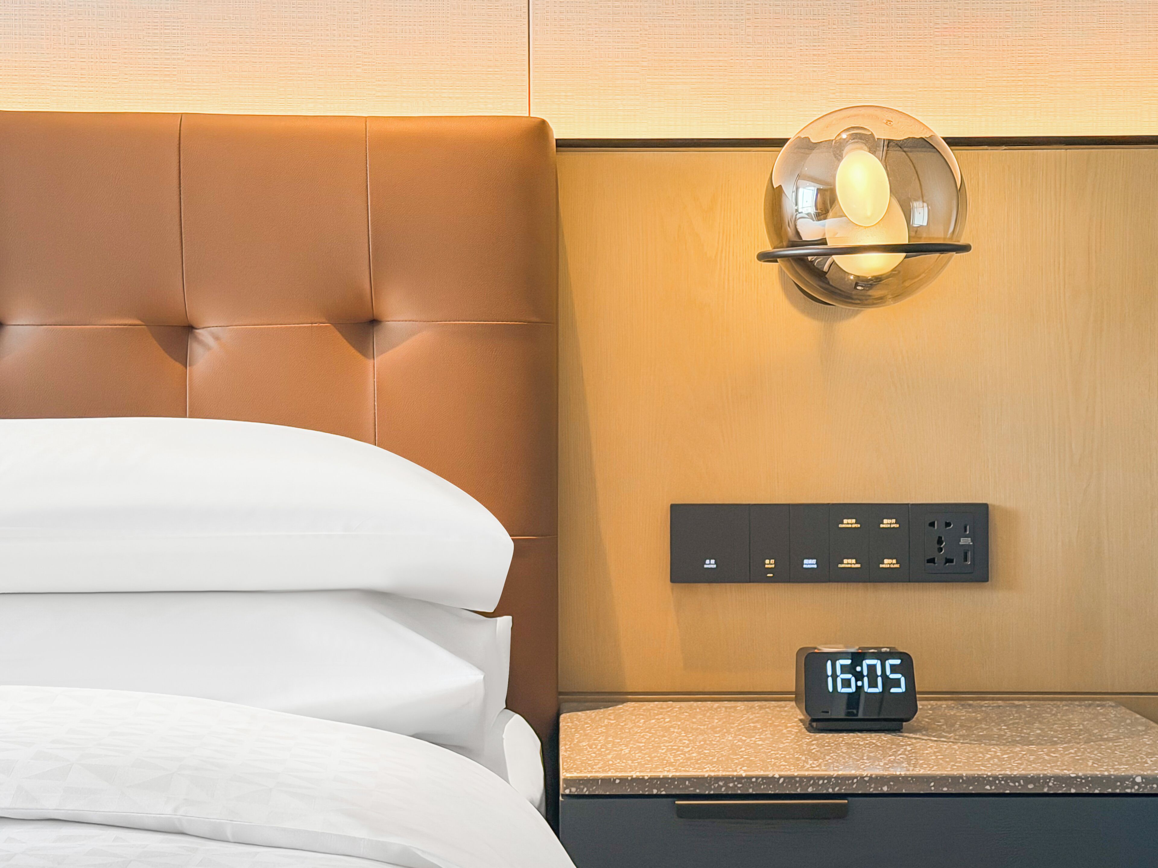 A hotel room with a tufted headboard, a digital clock, and a wall-mounted light fixture.