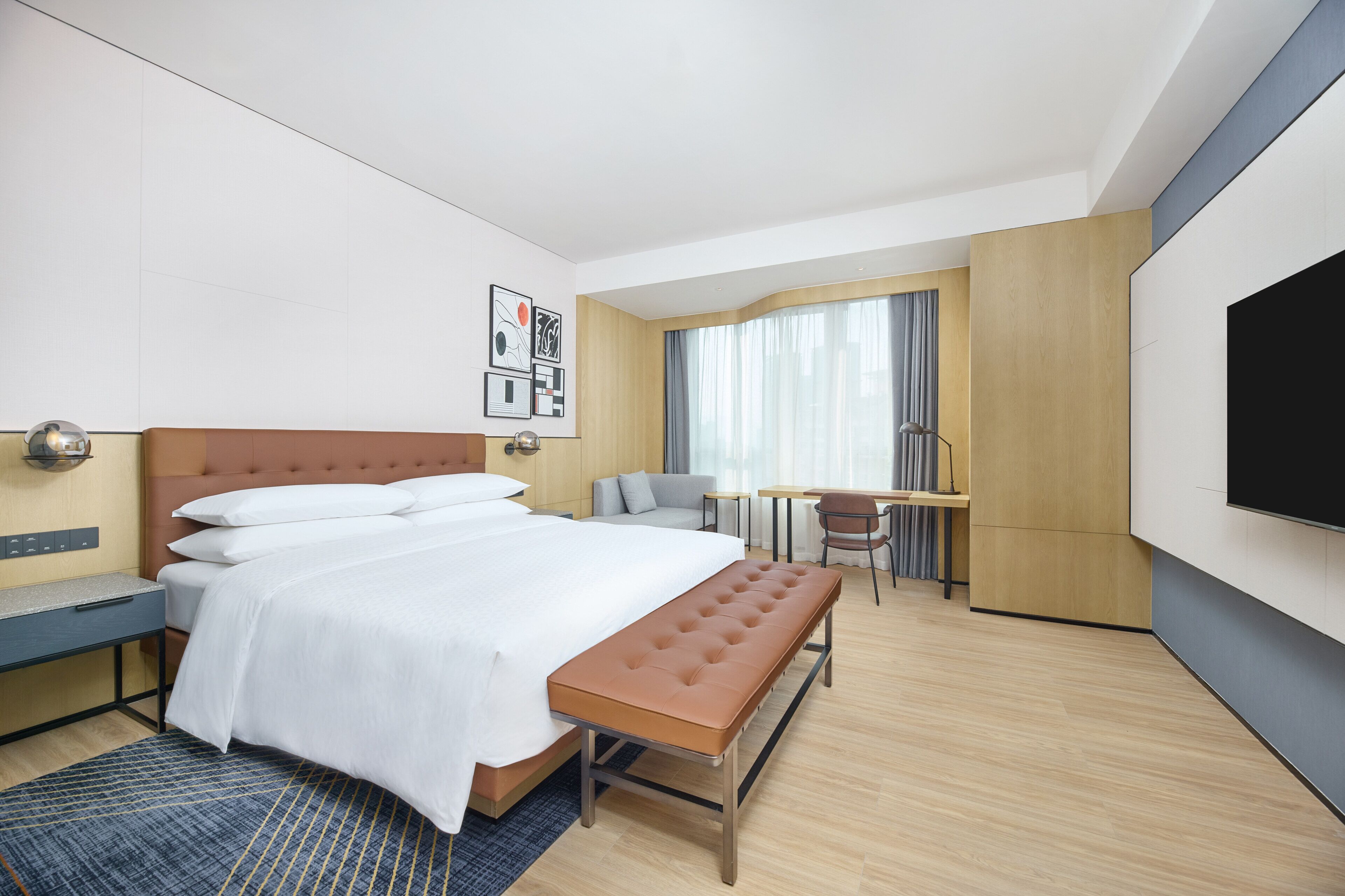 Ein Hotelzimmer mit einem Bett, einer Bank, einem Schreibtisch, einem Sessel, einem Fernseher und einem Fenster mit Vorhängen.