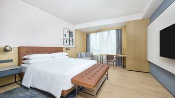 Ein Hotelzimmer mit einem Bett, einer Bank, einem Schreibtisch, einem Sessel, einem Fernseher und einem Fenster mit Vorhängen.