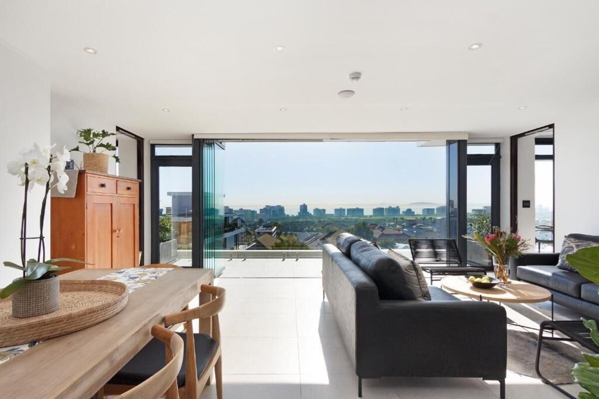 Un salon moderne avec un canapé, une table à manger et des chaises. De grandes fenêtres offrent une vue sur la ville.