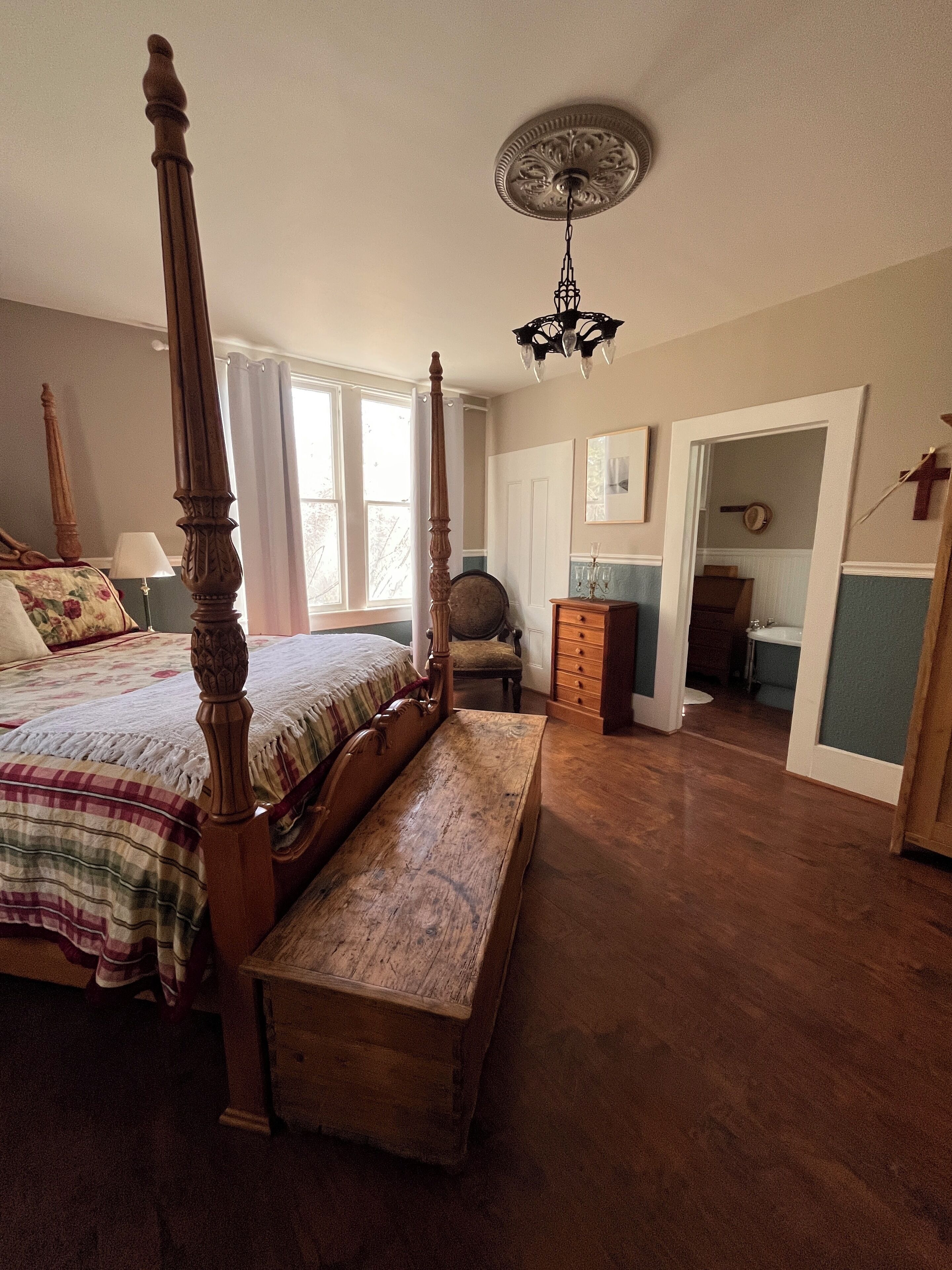 Un dormitorio con una cama con dosel, un banco de madera, un candelabro y vistas a un baño a través de una puerta abierta.
