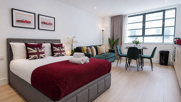 A modern hotel room with a grey bed, red throw, and a green sofa. There are two framed pictures on the wall, a window with curtains, and a small table with chairs.