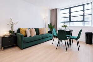 Una sala moderna con un sofá verde, una mesa de comedor con sillas, una lámpara, una planta en maceta y un ventanal grande.