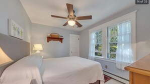 Une chambre à coucher comprenant un lit, un ventilateur de plafond, une fenêtre avec des rideaux, une étagère fixée au mur et une porte.