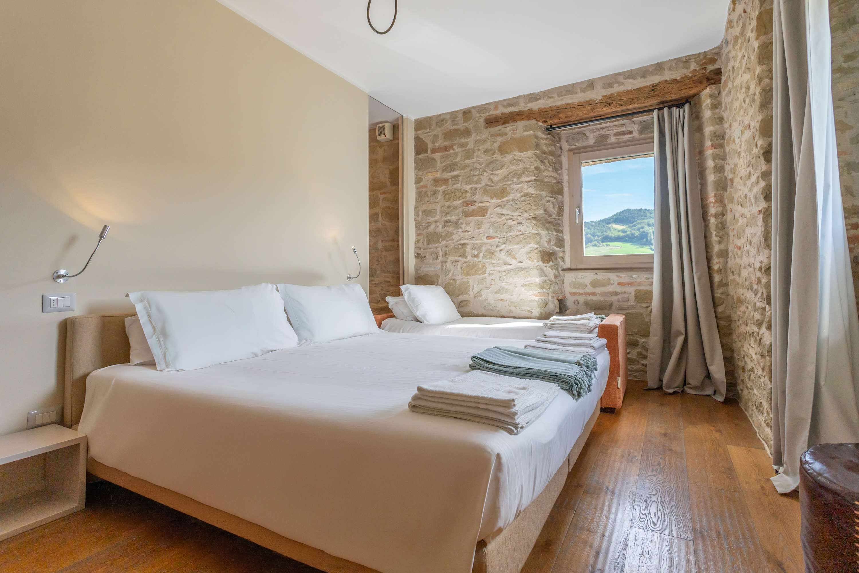 A bedroom with a large bed, a wooden floor, stone walls, a window with curtains, and a bedside table with a lamp.