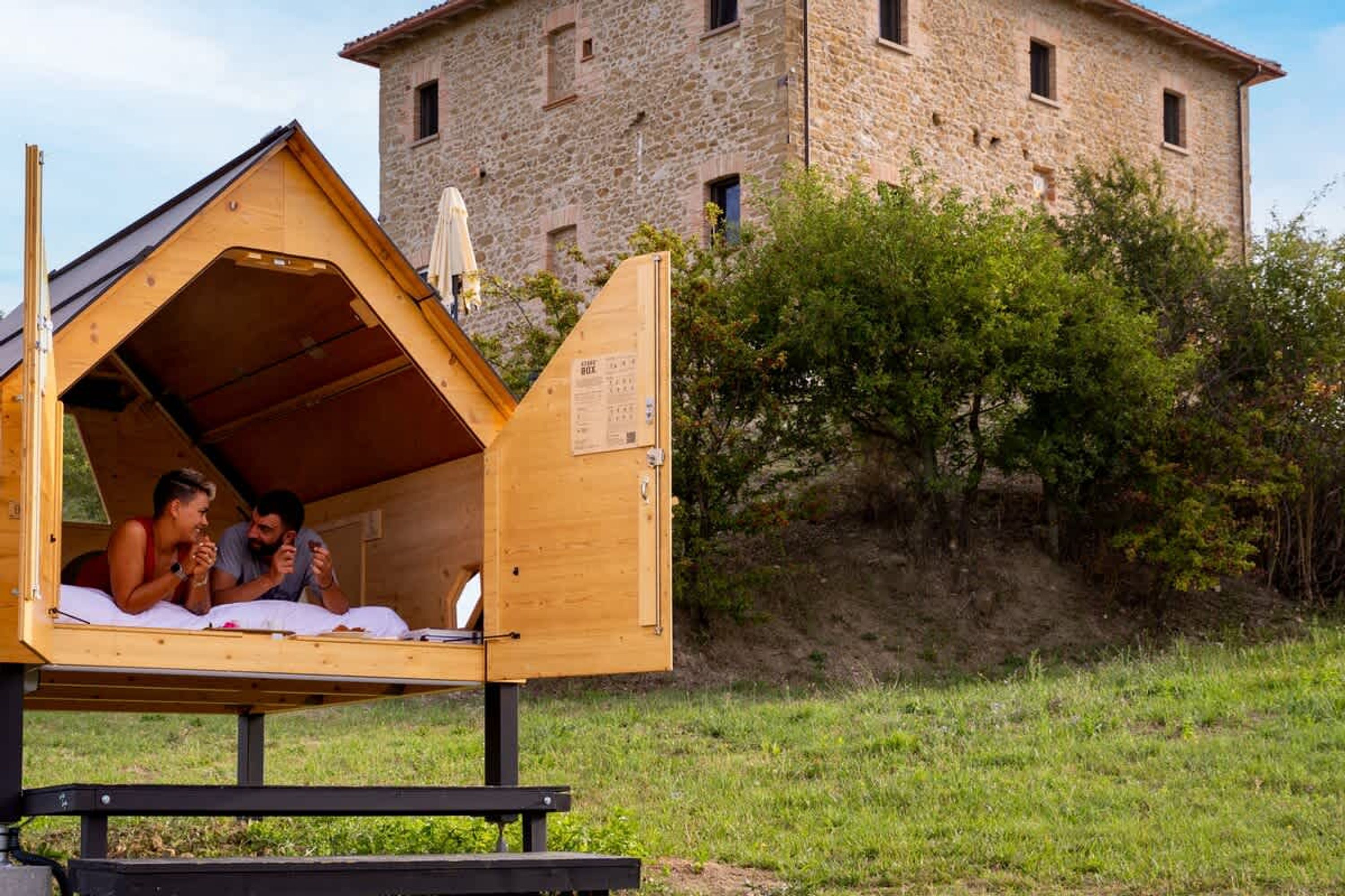 Une cabane en bois avec un toit à deux versants, une table de pique-nique devant et un bâtiment en pierre en arrière-plan.