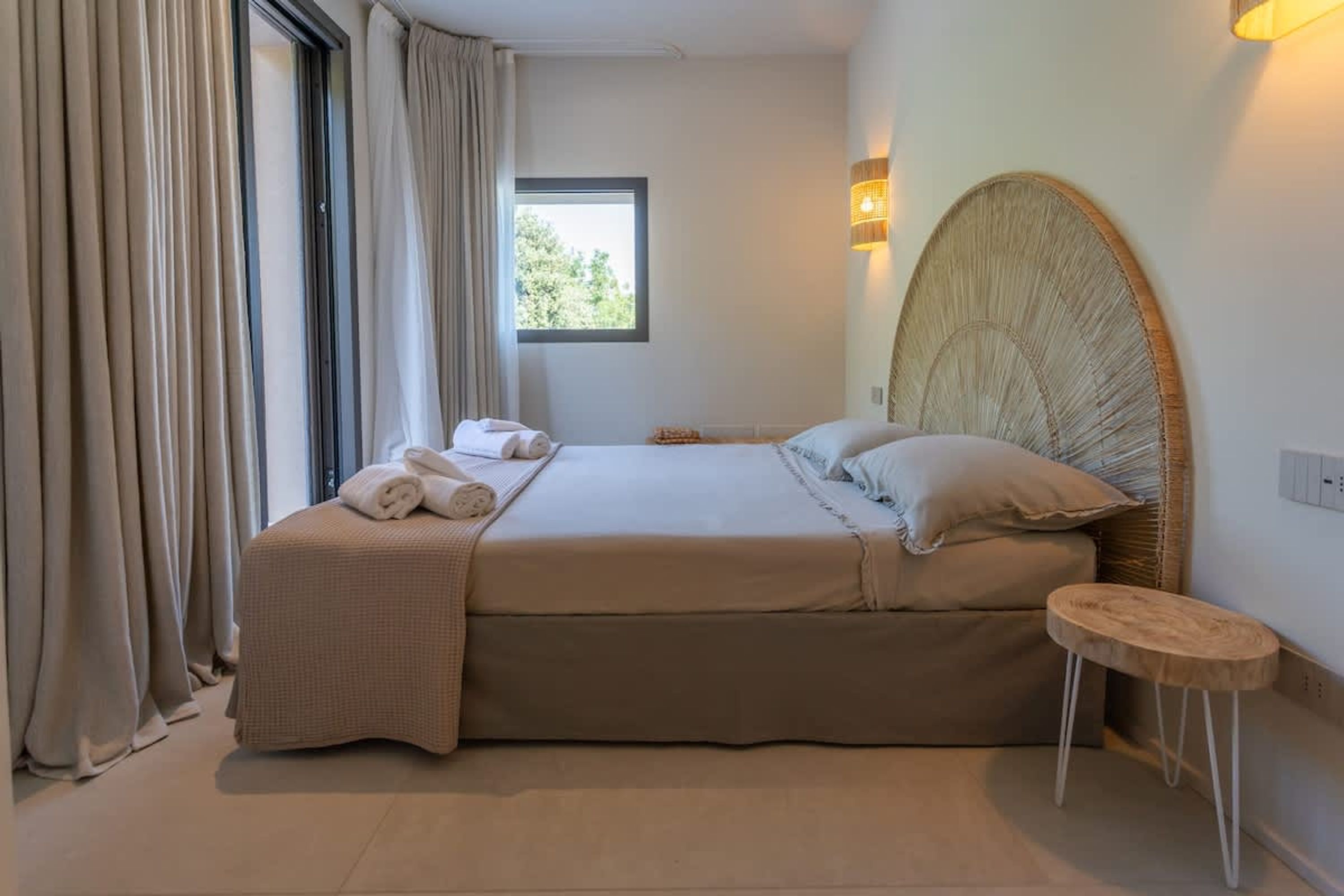 A bedroom with a large bed, a small stool, a window, and a wall-mounted lamp.