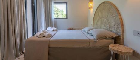 A bedroom with a large bed, a small stool, a window, and a wall-mounted lamp.