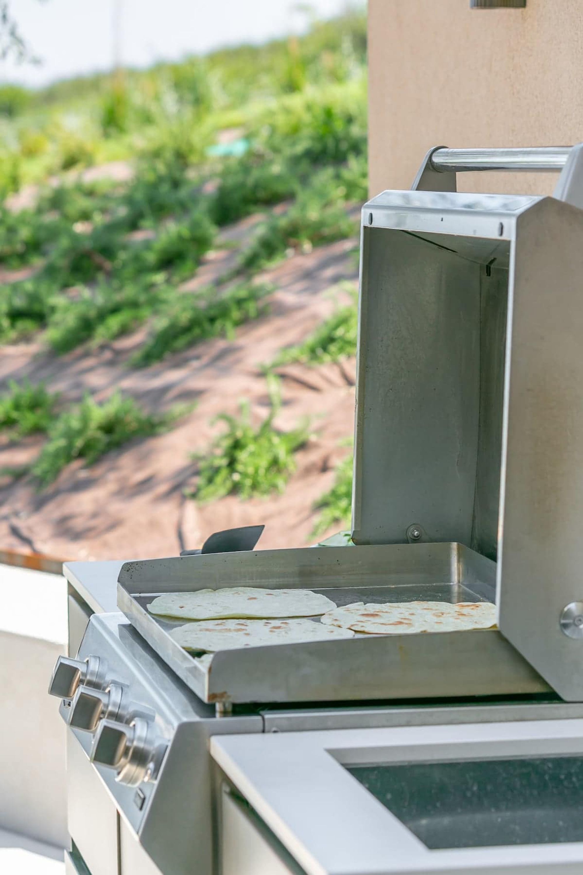 Un barbecue portable doté d’une structure métallique et d’une surface de cuisson plane, installé à l’extérieur, avec un arrière-plan flou de verdure.