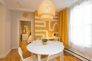 Un comedor con una mesa redonda blanca, sillas blancas, una lámpara colgante de fibras tejidas y un diseño abstracto amarillo y blanco en la pared.