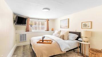 A bedroom with a bed, a TV mounted on the wall, a window with curtains, a bedside table with a lamp, and a small basket.