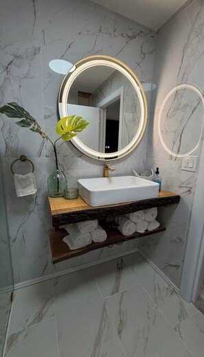 A bathroom with a marble backsplash, a round mirror with a gold frame, a white sink, and a shelf with rolled towels. - Oak & stone (Dalton)