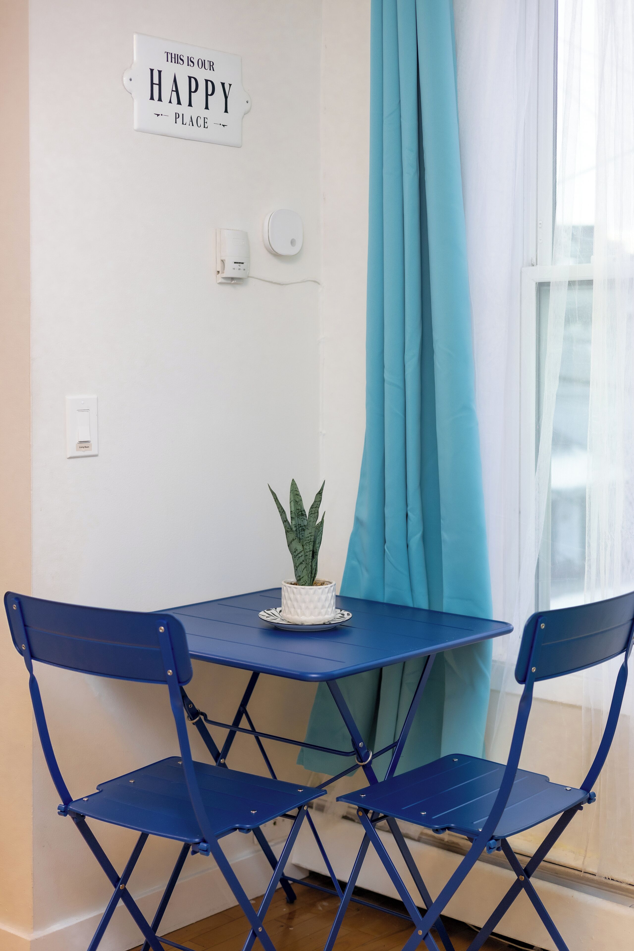 A small room with a blue table and chairs, a potted plant, a sign on the wall, and a window with curtains.