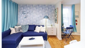 A living room with a blue sofa, white coffee table, and a desk with a chair. There is a patterned wallpaper on the wall and a window with curtains. - Quiet & Walkable - Stampede, BMO, Nightlife & Eats - Apartment (Calgary)