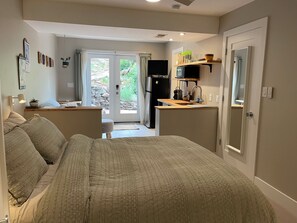 Une chambre avec un grand lit, une petite kitchenette équipée d’un réfrigérateur et une porte-fenêtre coulissante donnant sur un jardin.