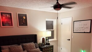 Quarto com uma cama, mesa de cabeceira com abajur, ventilador de teto e placa de boas-vindas.