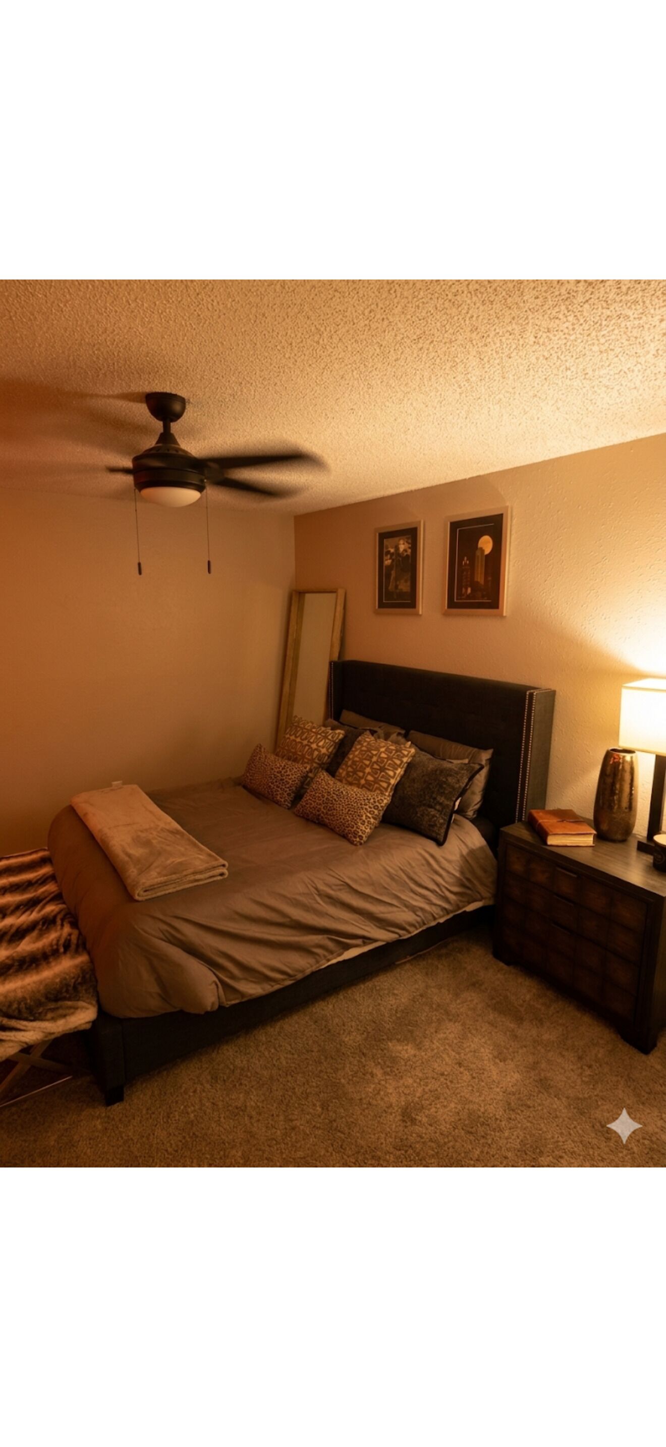 Een slaapkamer met een bed, een plafondventilator, een nachtkastje met een lamp en ingelijste foto's aan de muur.
