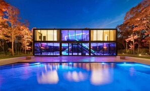 Un bâtiment moderne doté de grandes fenêtres en verre, une piscine au premier plan et des arbres aux feuilles d’automne en arrière-plan.
