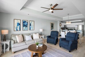 A living room with a sofa, armchairs, a coffee table, a ceiling fan, artwork on the wall, and a kitchen in the background. - Newly Updated Gulf Front 2-Bedroom Condo; Direct Ocean Views in Gulf Shores, AL (Gulf Shores)