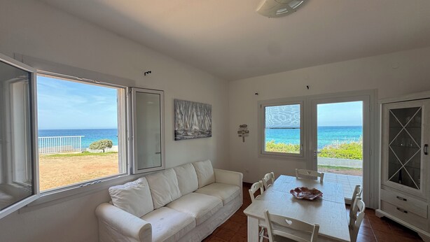 Un interior luminoso con un sofá blanco, una mesa de comedor y sillas. Amplias ventanas ofrecen vistas al océano.